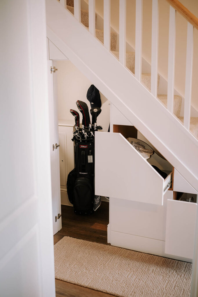 White under stairs storage with open cupboard door and pull-out drawer storing golf clubs in a hallway