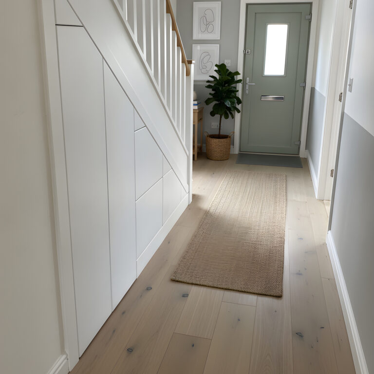 Design visualisation of white under-stairs storage in a bright hallway with a grey entrance door and woven runner.