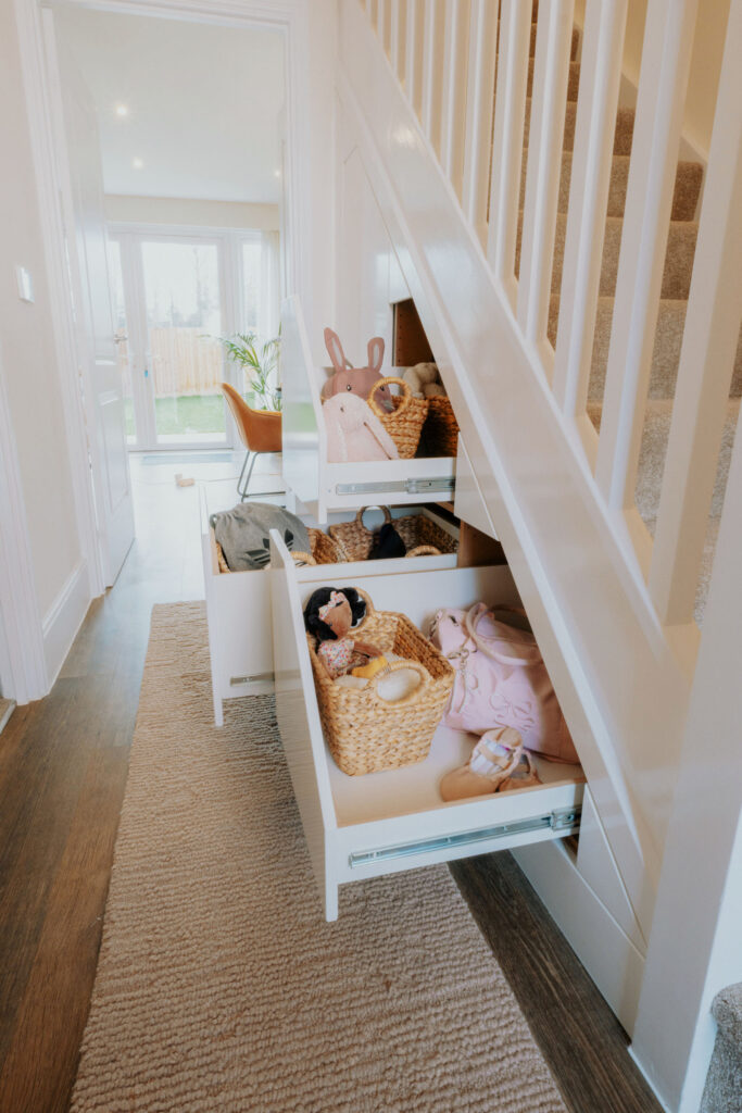 Open fitted under-stairs drawers holding shoes, bags and baskets in a family hallway.